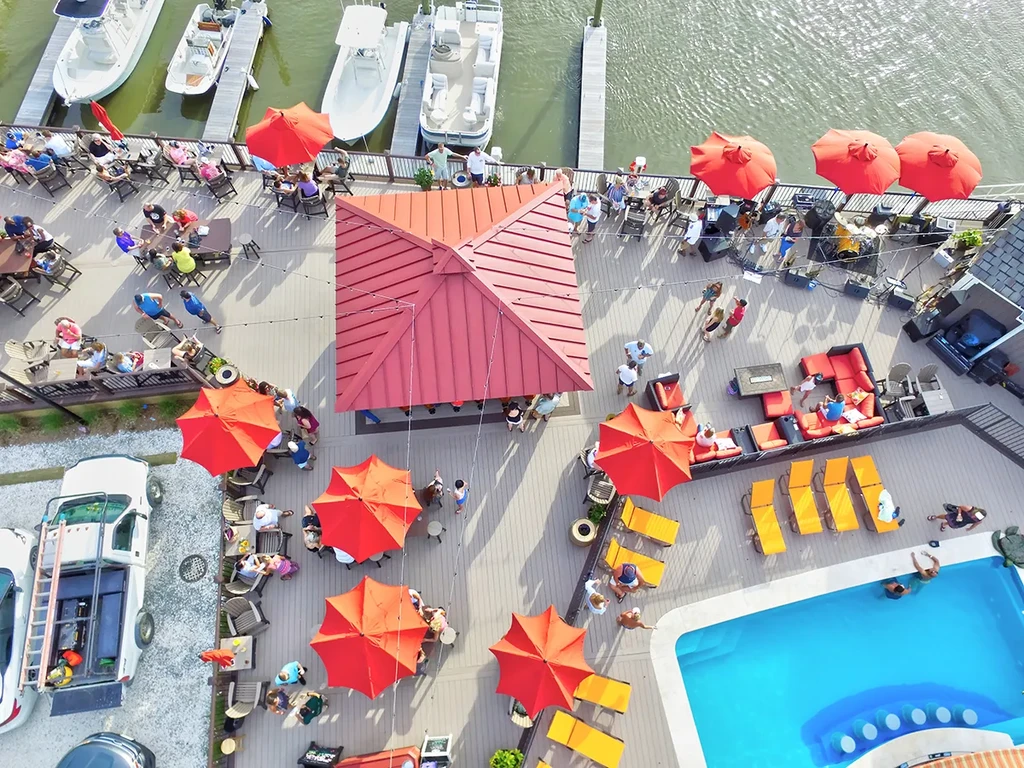 Aerial view: waterfront outdoor restaurant, pool, boats, red umbrellas, many people.