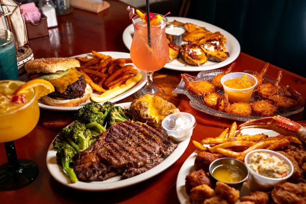 Table full of steak, burgers, fried seafood, and cocktails in a restaurant.