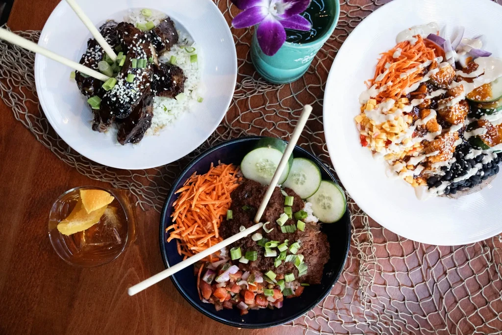 Top-down view of colorful food bowls, glazed ribs, and cocktails on a table.