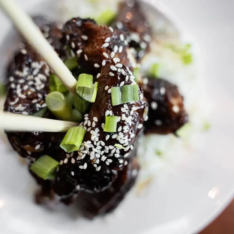 Chopsticks holding glazed meat with sesame seeds and green onions over rice.