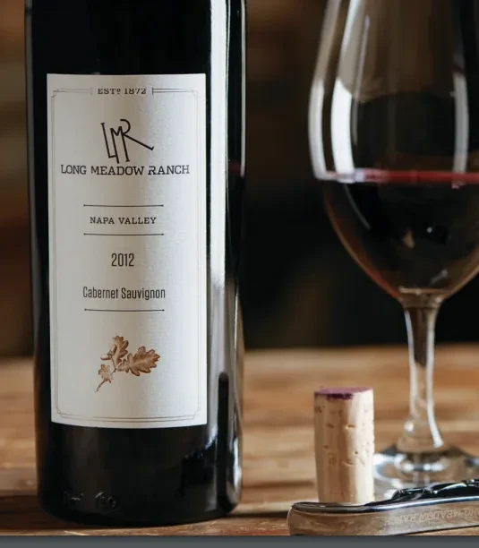 Long Meadow Ranch wine bottle, red wine glass, cork, corkscrew on wood.