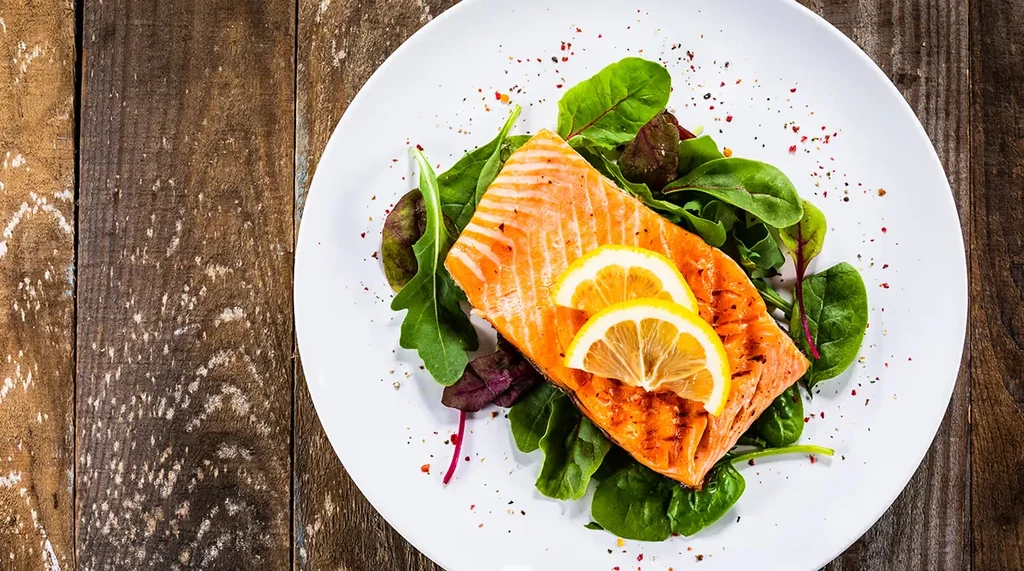 Grilled salmon with lemon and greens on white plate, wooden table.