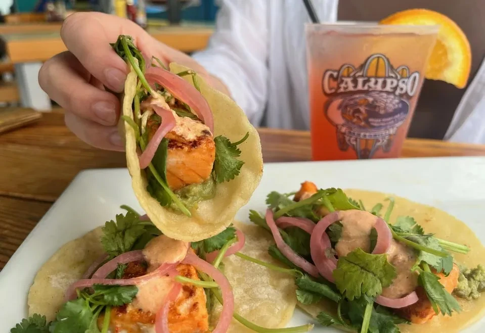 Grilled salmon taco held by a hand, two more on a plate.