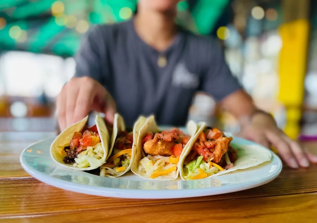 Three soft-shell tacos with fish/chicken, lettuce, cheese, salsa; person reaching.