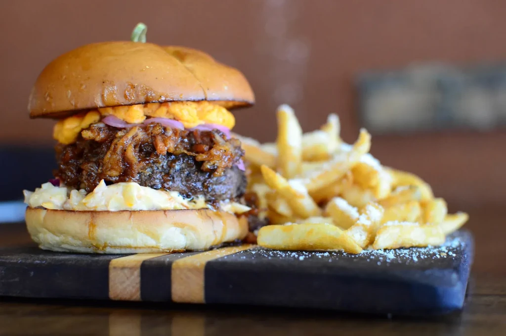 Close-up of a stacked burger and cheesy fries on a dark board.