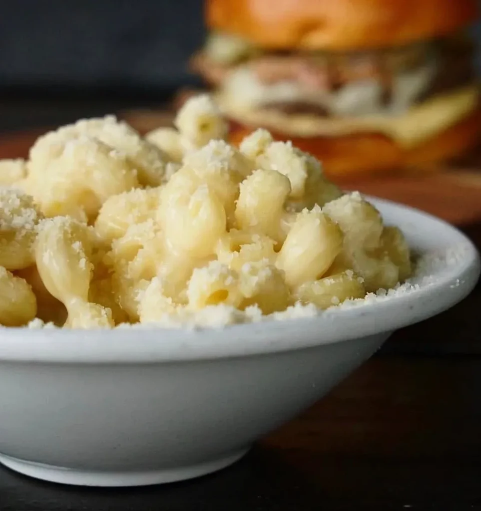White bowl of macaroni and cheese with Parmesan, blurred burger behind.