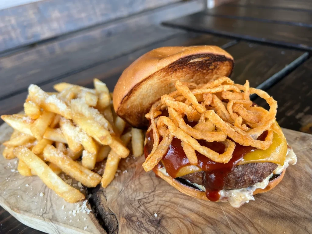 Burger with melted cheese, onion strings, and seasoned fries on wood.