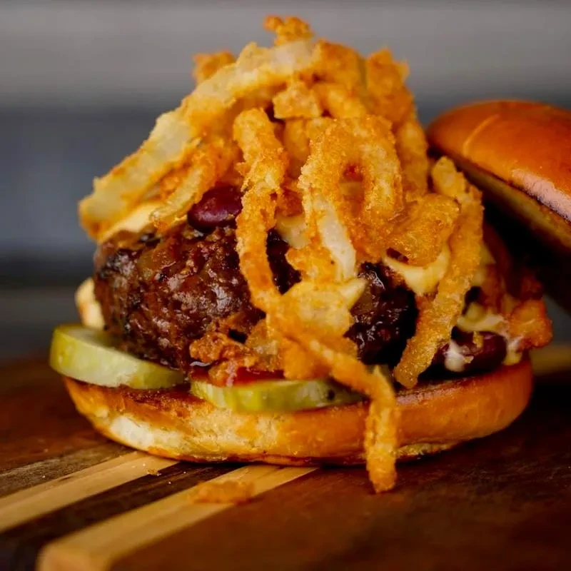 Stacked gourmet burger with beef, chili, pickles, and fried onion rings.