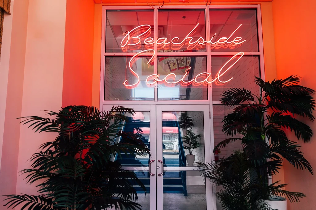 Neon "Beachside Social" sign illuminates a building entrance with palm trees.