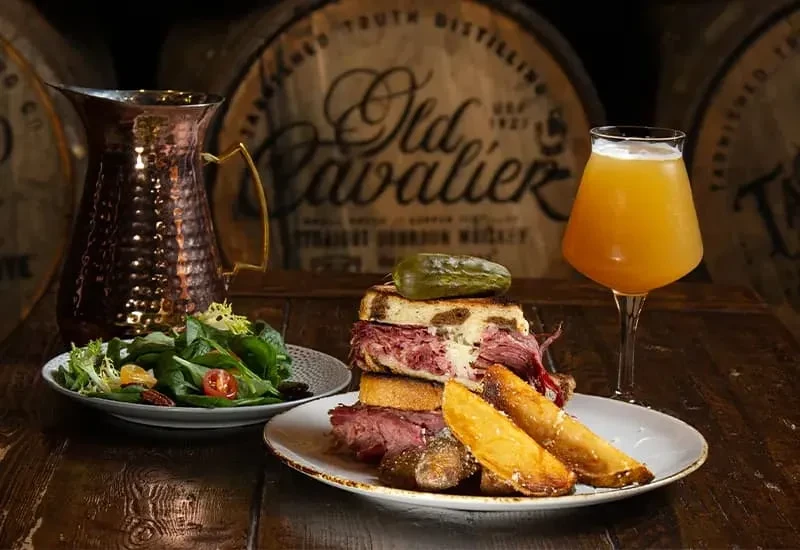 Tall sandwich, potato wedges, salad, and beer at a distillery.