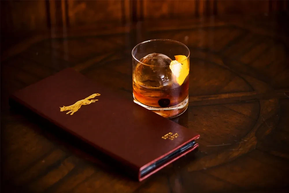 Amber cocktail, spherical ice, orange peel, and fox-logo menu on wood.