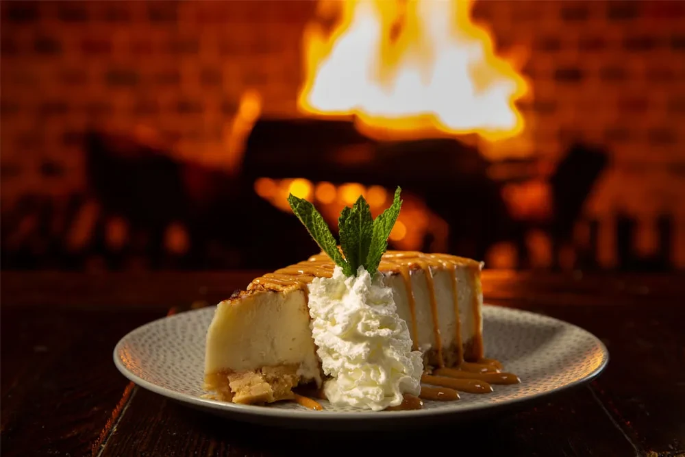 Cheesecake slice with whipped cream, caramel, and mint by a fireplace.