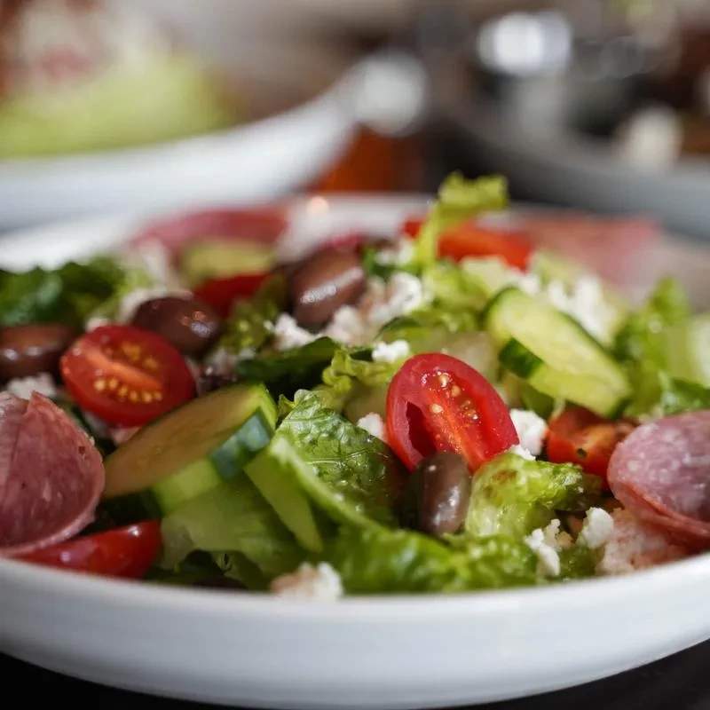 Salad in white bowl with lettuce, tomatoes, olives, cheese, and salami.