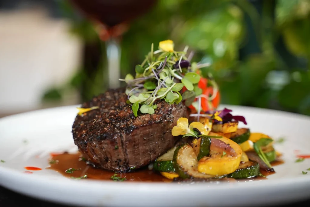 Seared meat, microgreens, roasted vegetables, and dark sauce on a white plate.