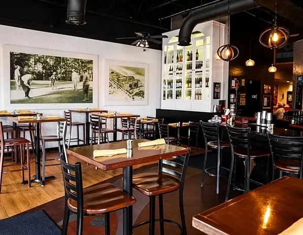 Restaurant interior with high-top tables, bar area, and illuminated bottle shelves.