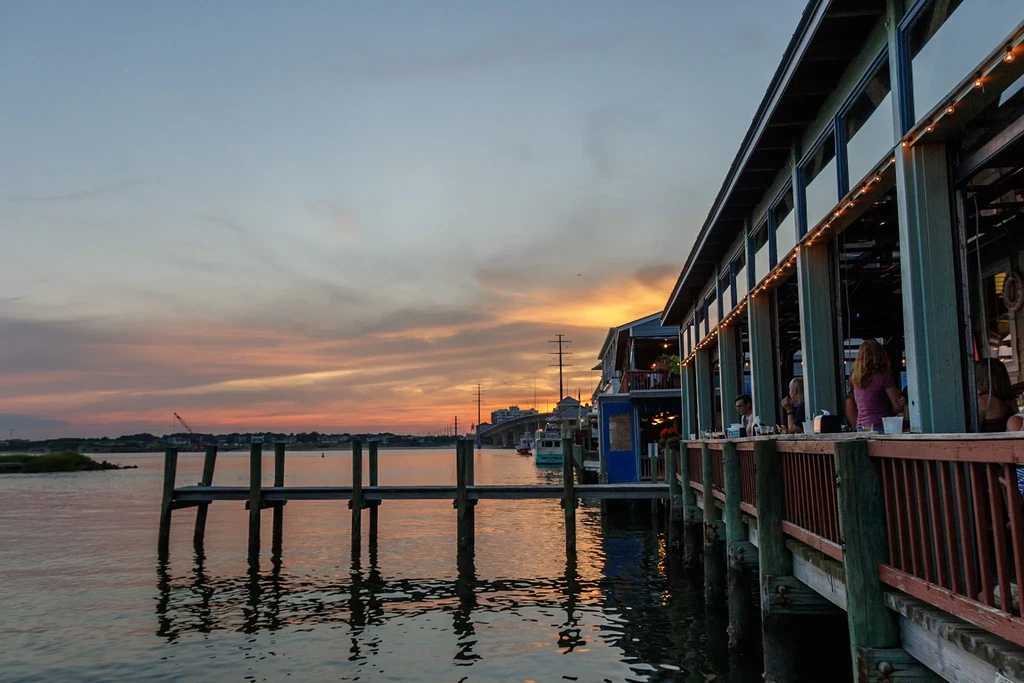 Sunset over water with restaurant, pier, and distant bridge.