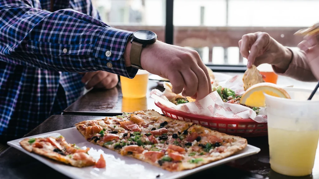 Close-up of hands sharing flatbread pizza, nachos, and beverages.