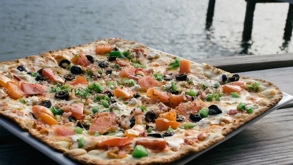 Pizza with olives, peppers, feta, and cured fish by the water.