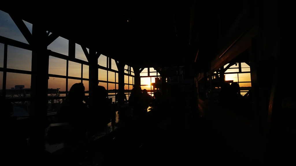 Restaurant interior with silhouetted patrons watching sunset over water.