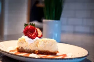 Cheesecake slice with graham crust, strawberries, whipped cream, and red sauce.