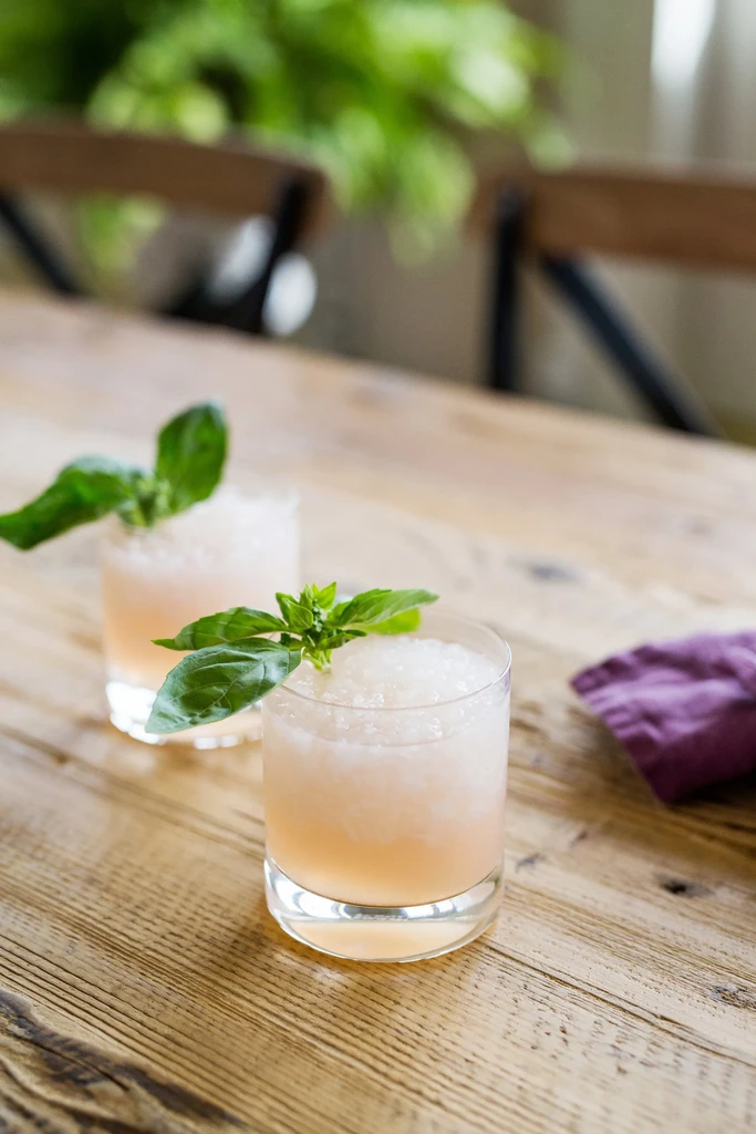 Two slushy peachy-pink drinks with basil on a rustic wooden table.