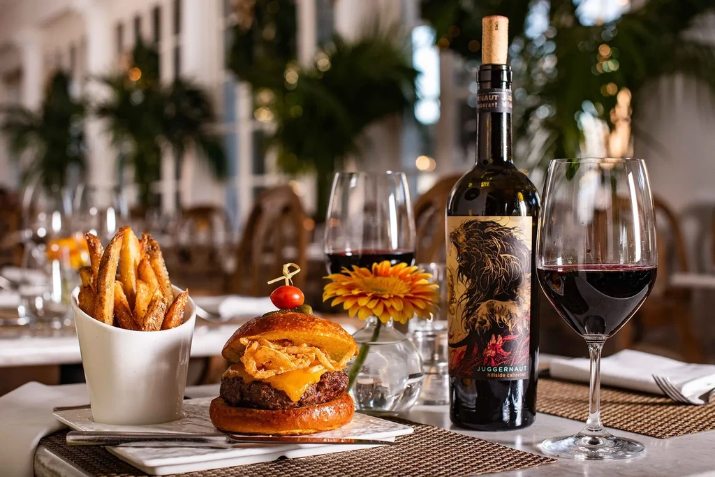 Restaurant table with a gourmet burger, fries, red wine, and daisy.