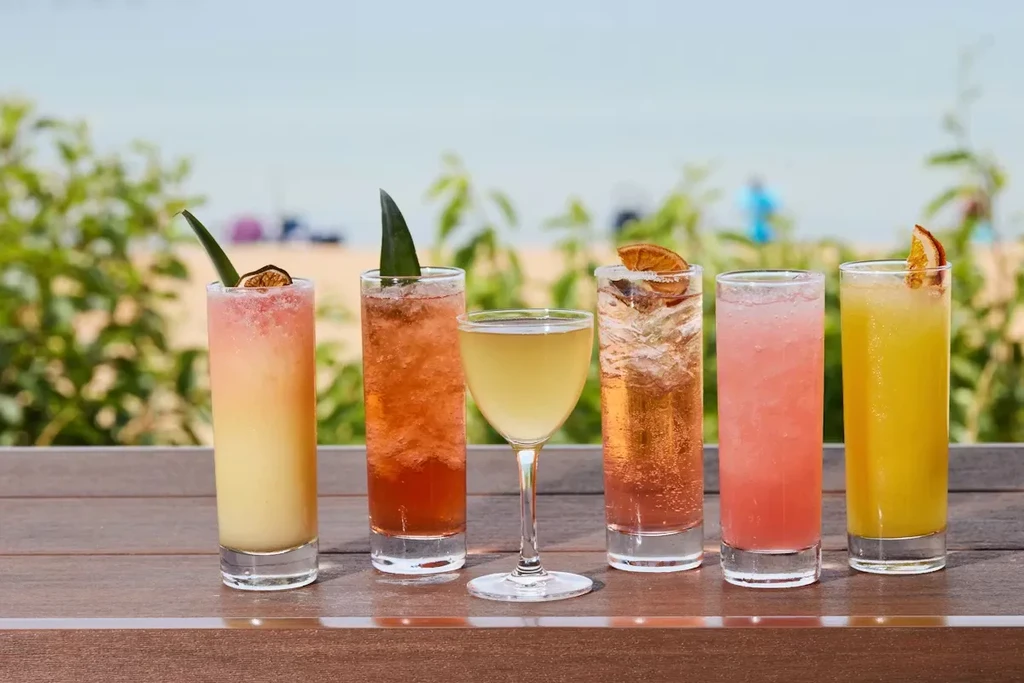 Six assorted cocktails on a wooden surface, with a beach backdrop.