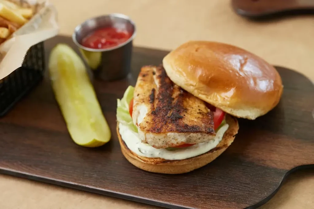 Seared fish sandwich, pickle, fries, and ketchup on a paddle board.