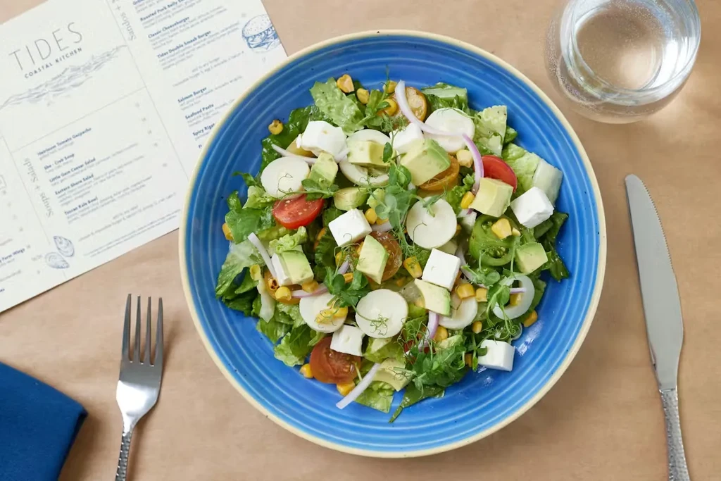 Vibrant salad in blue bowl, with menu, fork, and water.