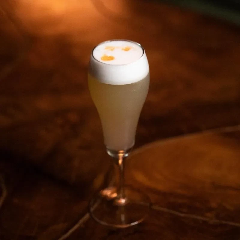 Pale cocktail in tall glass with white foam and orange dots.