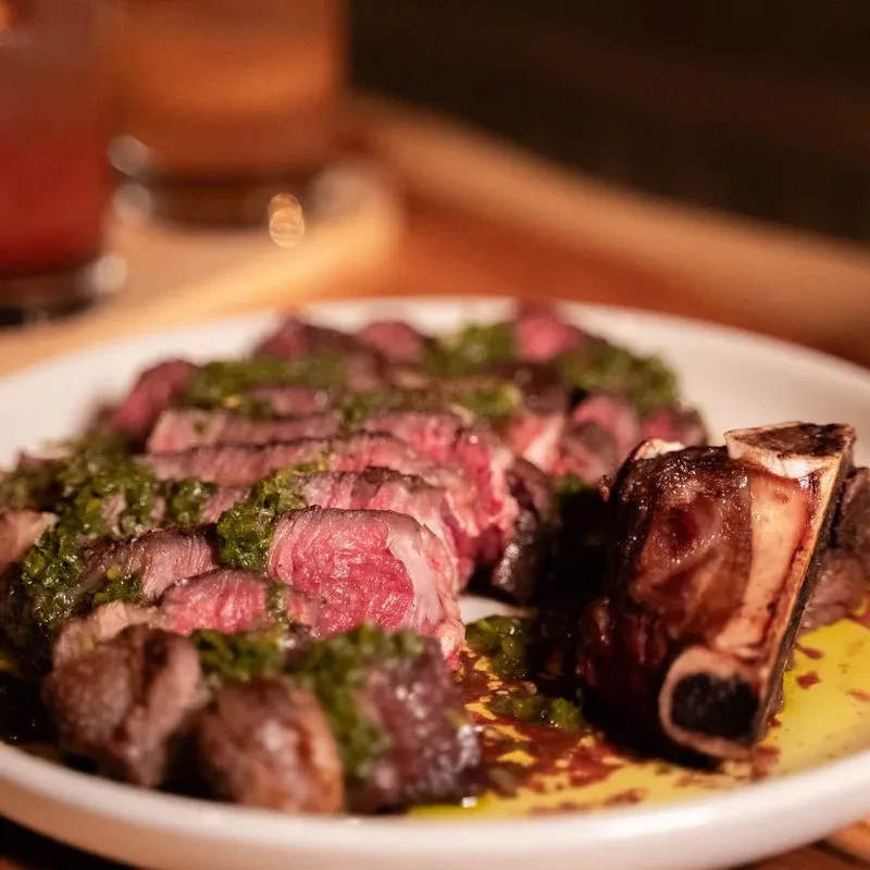 Plate of sliced steak, green sauce, and a bone-in piece.