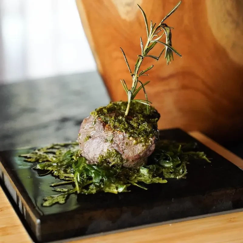 Thick cut meat with green herb sauce, rosemary, and leafy greens.