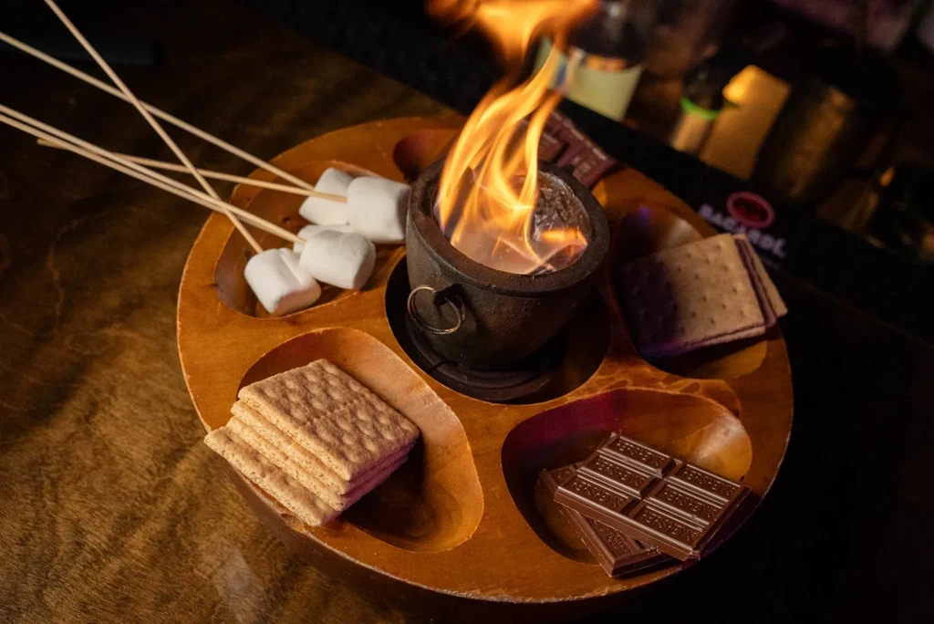 Overhead s'mores kit on wooden tray with fire, marshmallows, chocolate, crackers.