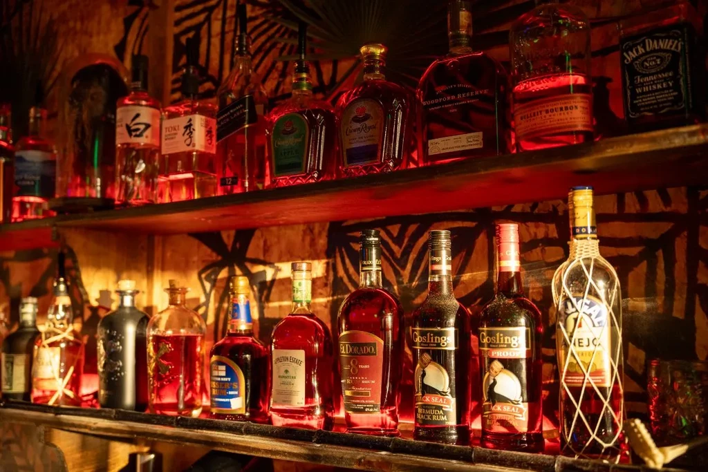 Warmly lit bar shelf displaying whiskey and rum bottles, patterned background.