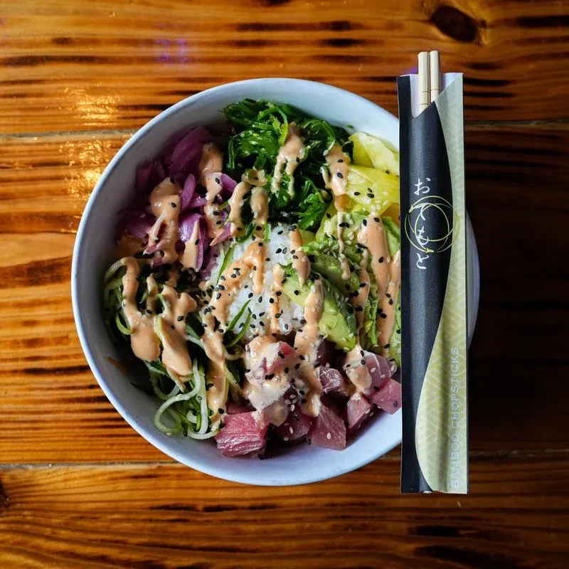 Overhead poke bowl with tuna, avocado, seaweed, chopsticks on wooden table.