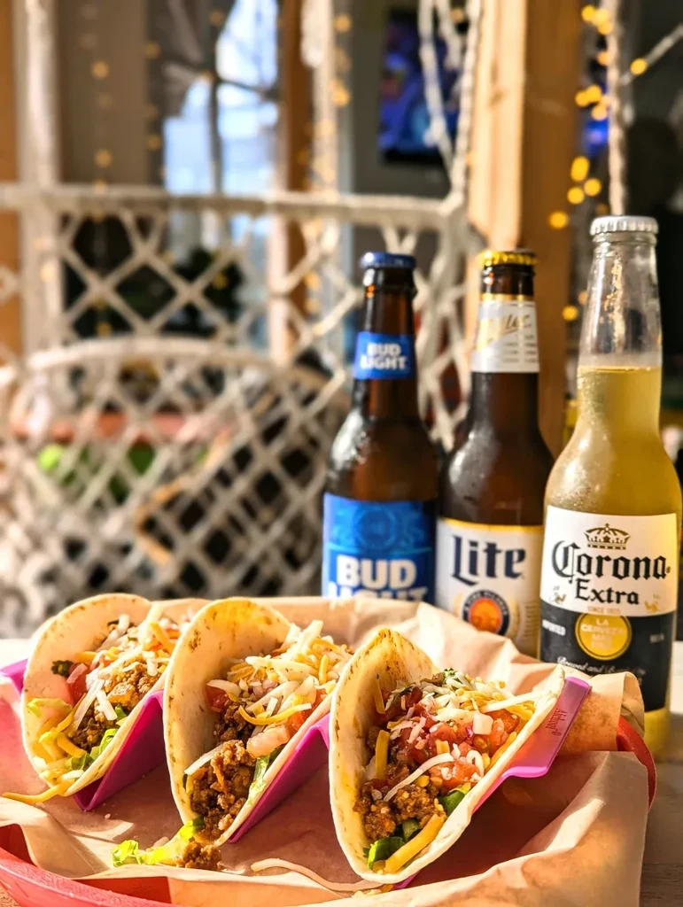 Three meat tacos in pink holders with assorted beer bottles.