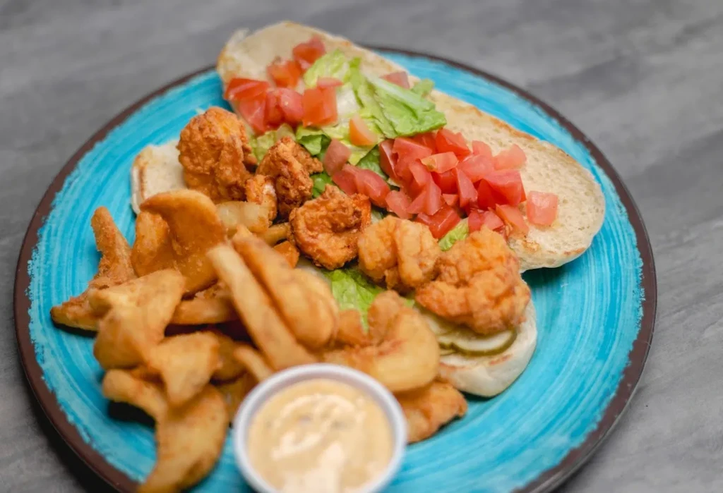 Fried shrimp Po'boy with wedge fries and sauce on a blue plate.