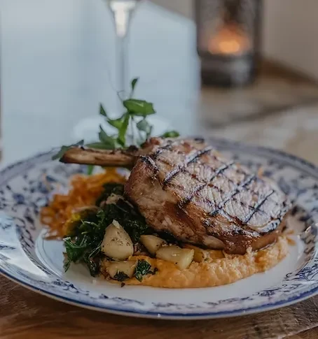 Grilled pork chop, creamy purée, greens, garnished with herbs on a patterned plate.