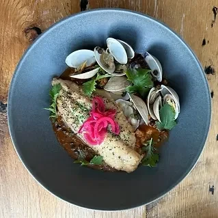 Pan-seared fish with clams, pickled onions, and herbs in a dark bowl.