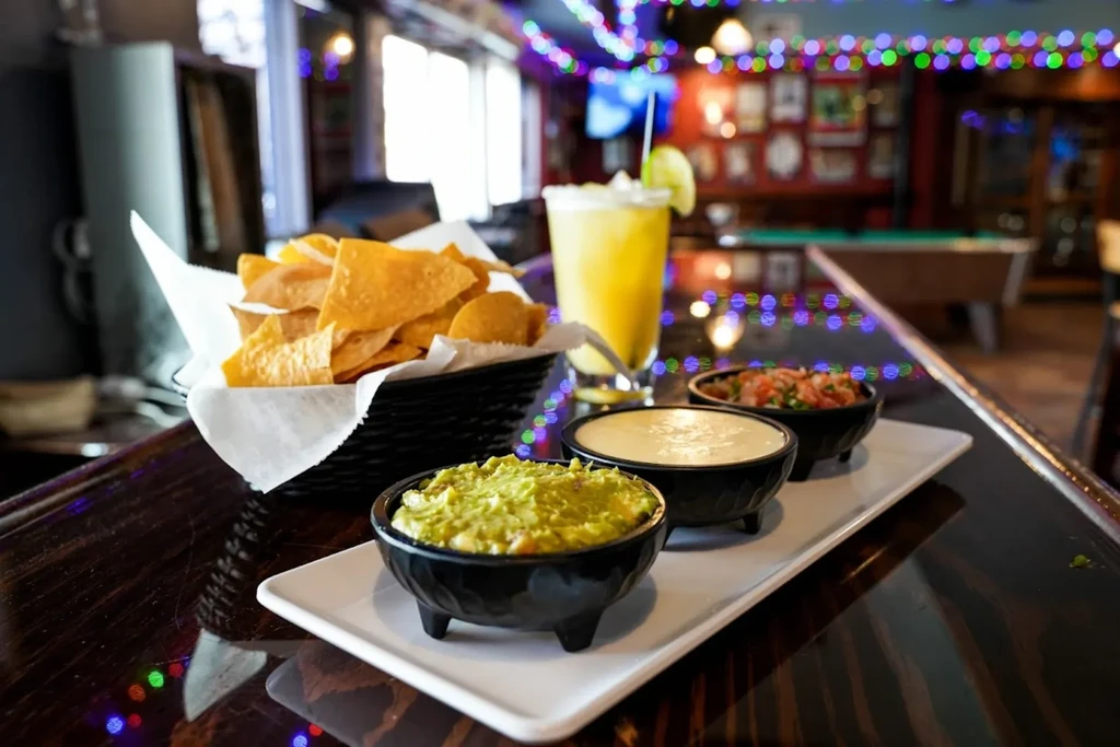 Tortilla chips, three dips, and margarita on a festive bar counter.