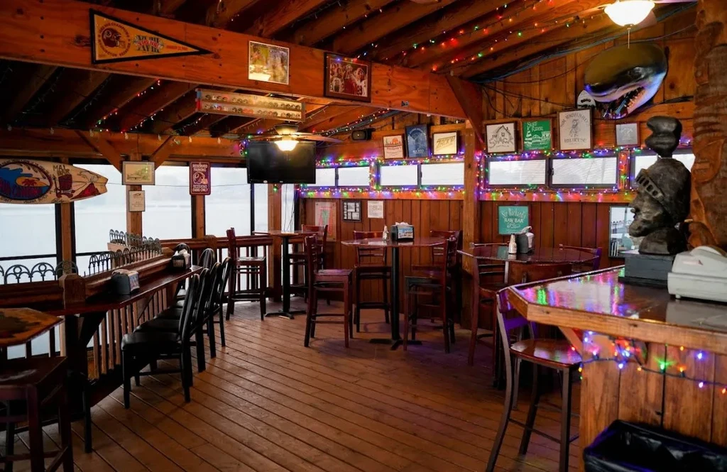 Empty, rustic restaurant with warm wood, string lights, and waterfront view.