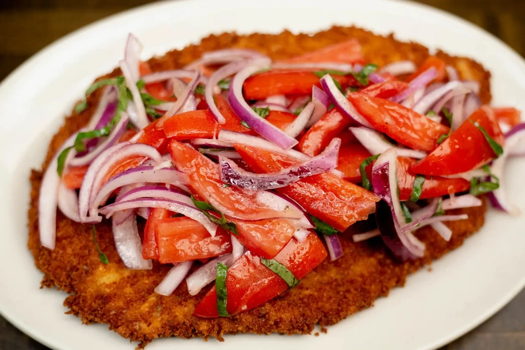 Crispy golden-brown breaded cutlet topped with tomato, red onion, and herbs.