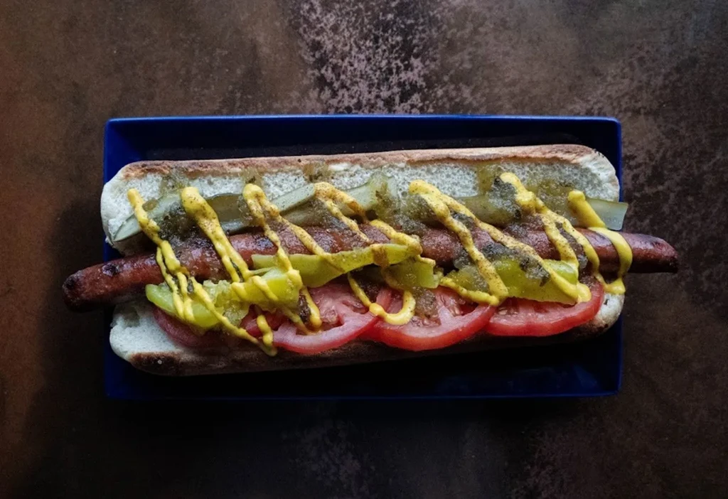 Grilled hot dog in a bun, topped with tomato, relish, and mustard.
