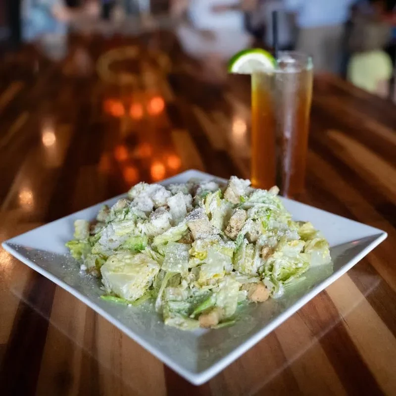 Caesar salad and amber drink with lime on a wooden table.