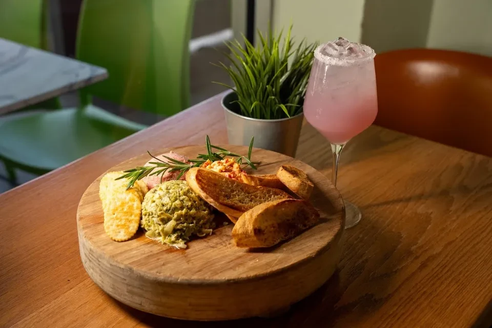 Wooden platter with appetizer spreads, crackers, baguette, and pink cocktail.