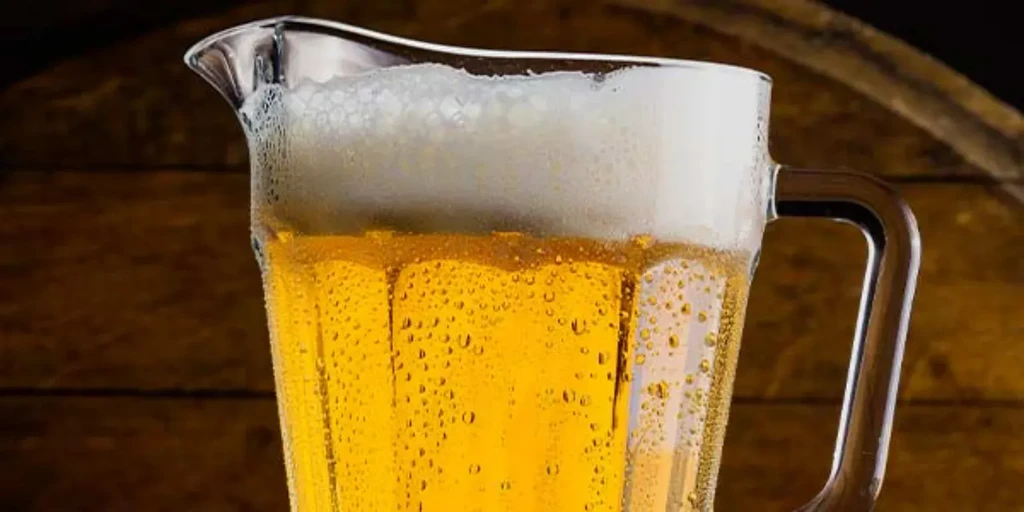 Cold glass pitcher of golden beer with frothy head on wood.