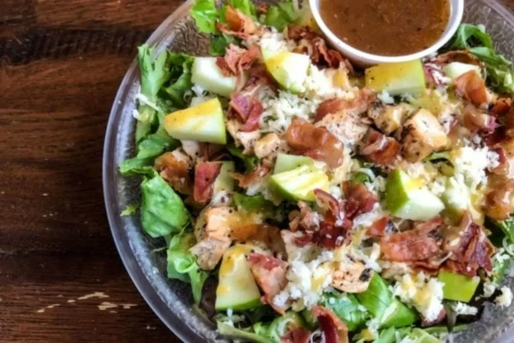 Chicken, bacon, and apple salad with dressing in a clear bowl.