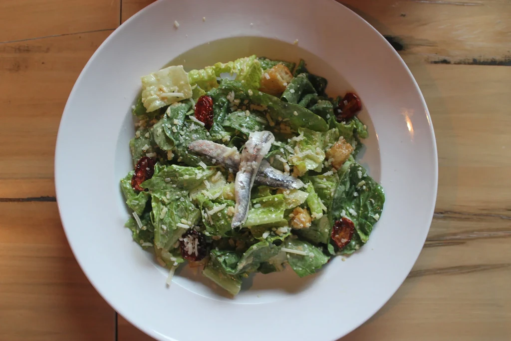 Overhead Caesar salad with crossed anchovy fillets, cheese, croutons, and dried fruit.
