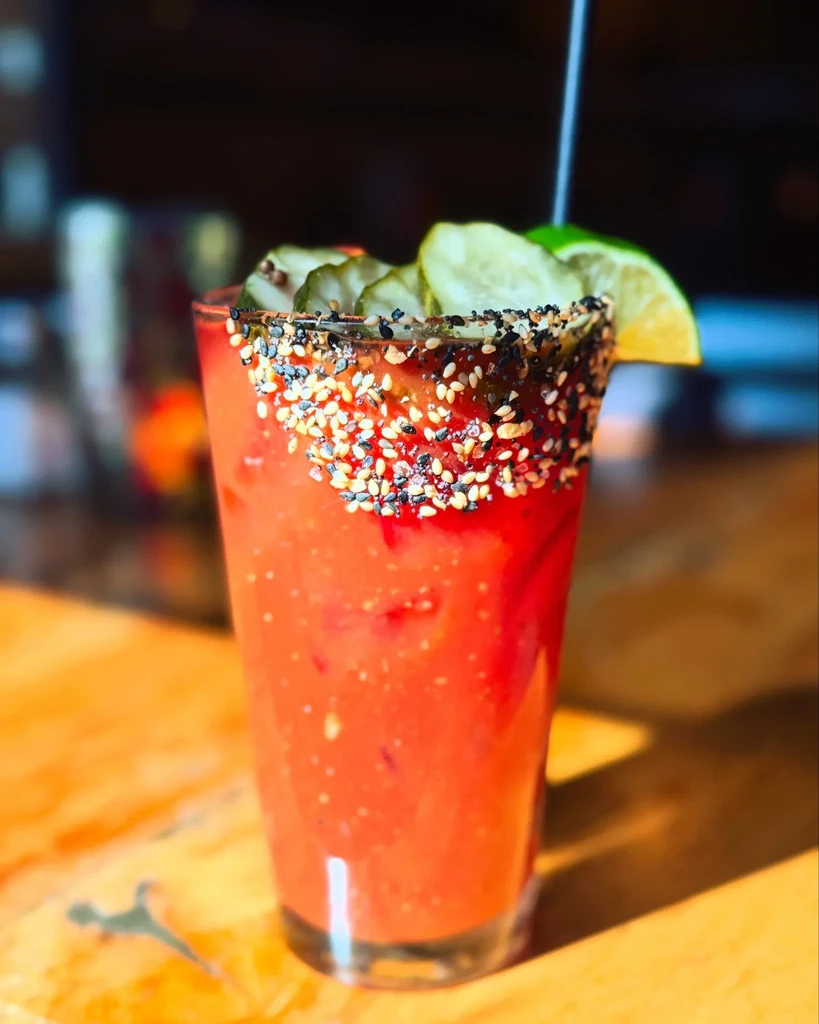 Reddish-orange cocktail with everything bagel rim, pickles, and lime.
