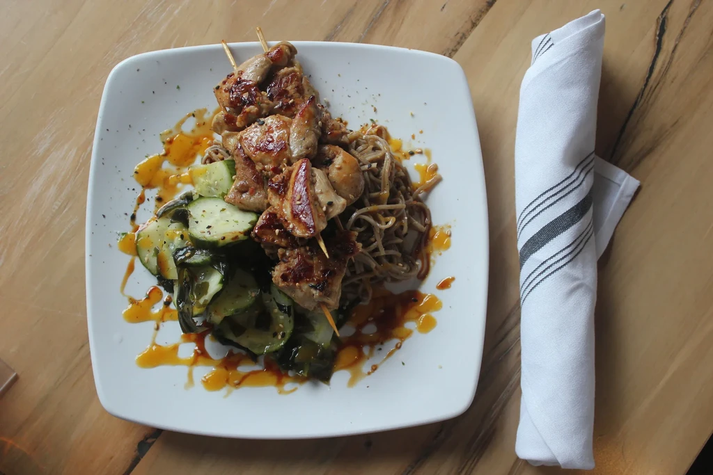 Overhead view of grilled chicken skewers with noodles, cucumbers, sauce, and napkin.